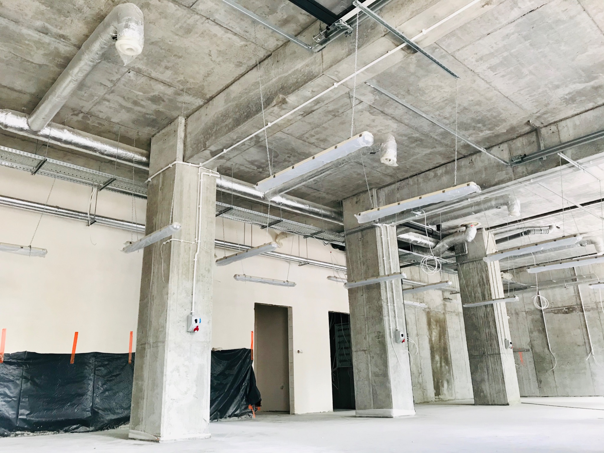Wnętrze budynku komercyjnego w trakcie instalacji systemu wentylacji: widoczne surowe betonowe stropy z kanałami wentylacyjnymi w folii ochronnej, podwieszone lampy tymczasowe i słupy konstrukcyjne...