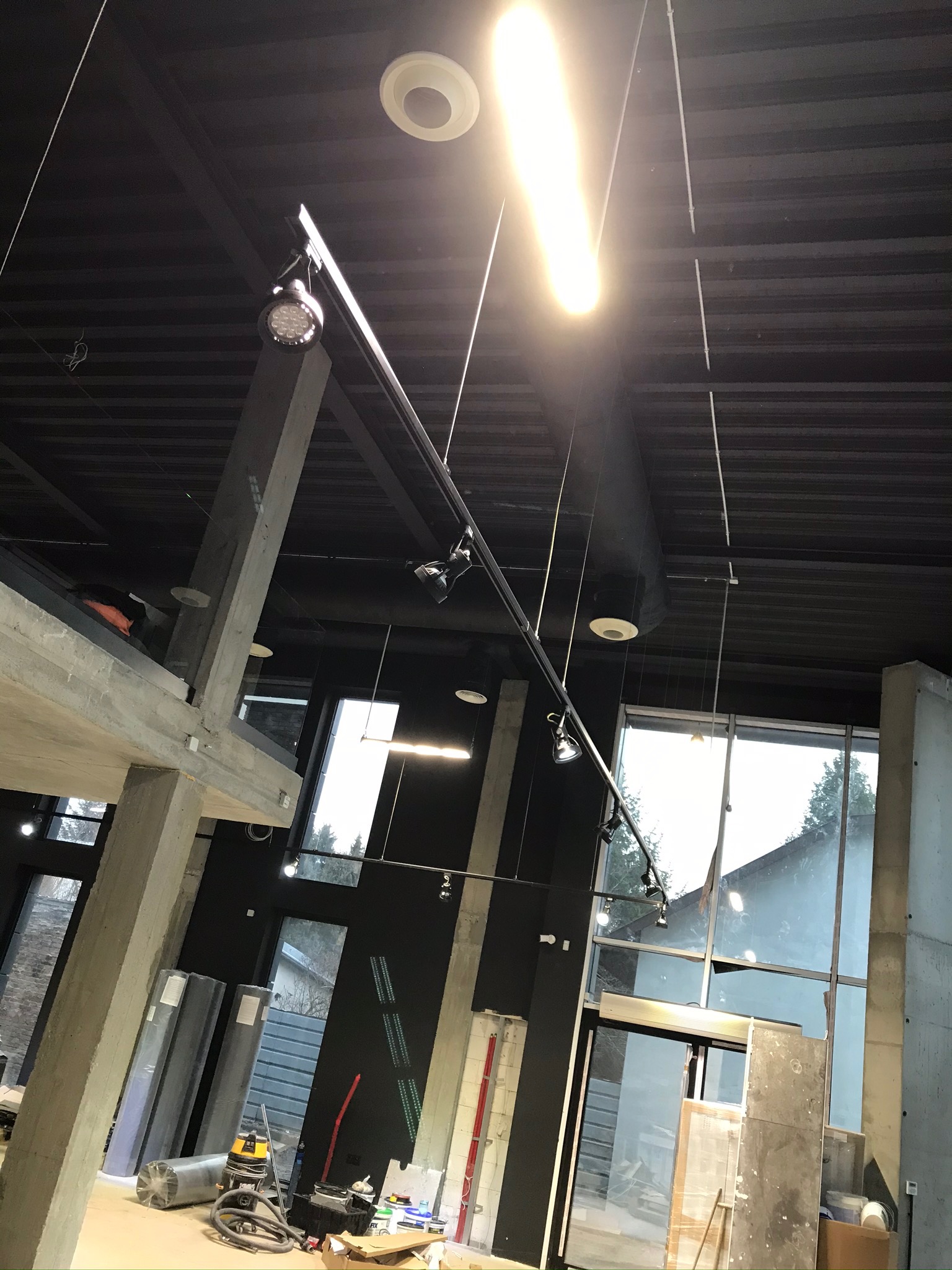 Wnętrze budynku komercyjnego w trakcie montażu instalacji wentylacyjnej, widoczne elementy systemu podwieszone pod sufitem, surowy beton, duże okna i materiały budowlane.