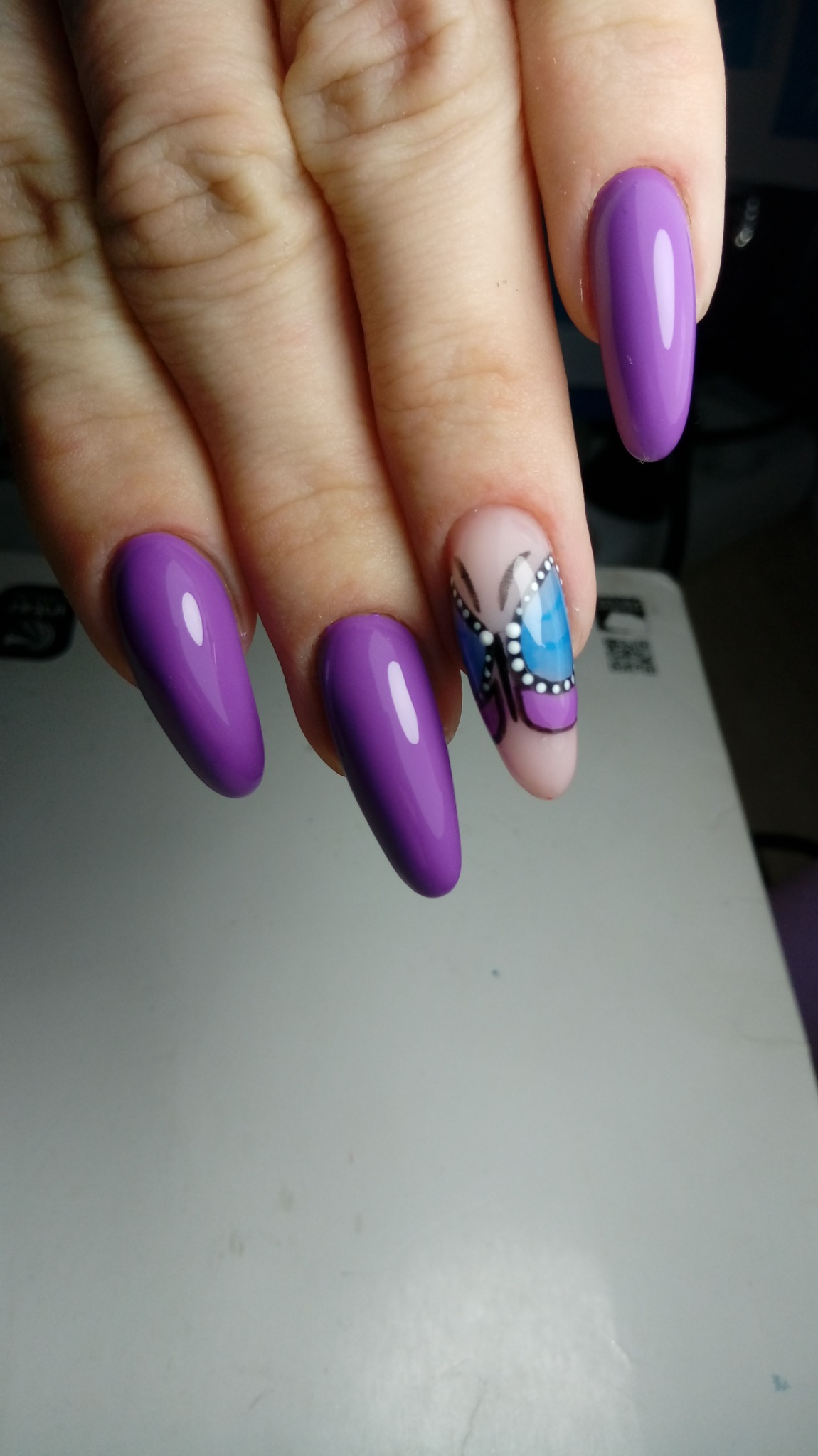 Dłoń z fioletowymi, błyszczącymi paznokciami w kształcie migdała; jeden paznokieć z motywem motyla w odcieniach błękitu i fioletu.
