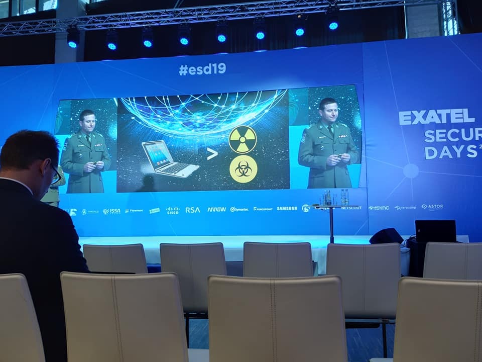 Prelegent w mundurze na konferencji bezpieczeństwa danych #esd19, na ekranie laptop zestawiony ze znakiem promieniowania i biohazardu, widoczne logotypy sponsorów.