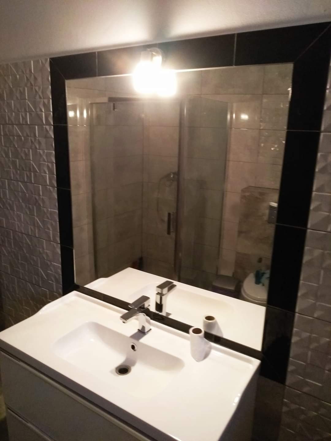 Biała umywalka z szafką, chromowana bateria i dekoracyjny wazonik odbijają się w lustrze z czarną ramą; w tle kabina prysznicowa i strukturalna szara ściana.