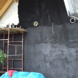 Ściana w trakcie prac przygotowawczych do wykończenia, częściowo pokryta ciemną warstwą, widoczne otwory na instalacje, rusztowanie, drewniana konstrukcja dachu i fragment sofy w pomieszczeniu.