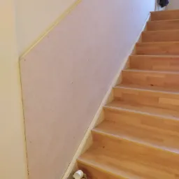 Schody drewniane z zamontowaną płytą na ścianie w trakcie prac wykończeniowych, widoczne ślady po wkrętach i małe pudło z kubkiem na schodach.