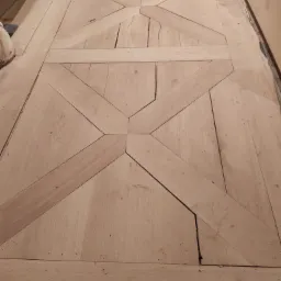 Układanie parkietu w geometryczny wzór krzyża z jasnego drewna, widoczne niedociągnięcia i szczeliny między klepkami.