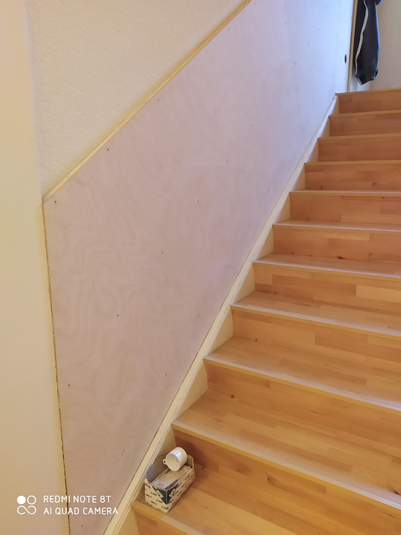 Ściana przy schodach wykończona płytą, widoczne drobne niedoskonałości montażu i pudełko z kubkiem na schodach.