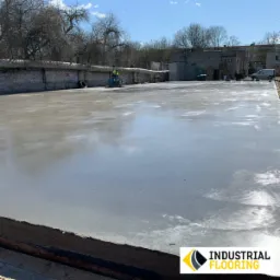 Gładka, szara wylewka betonowa na zewnątrz budynku, obrabiana przez dwóch pracowników za pomocą maszyny szlifierskiej; widoczne budynki w tle i błękitne niebo.