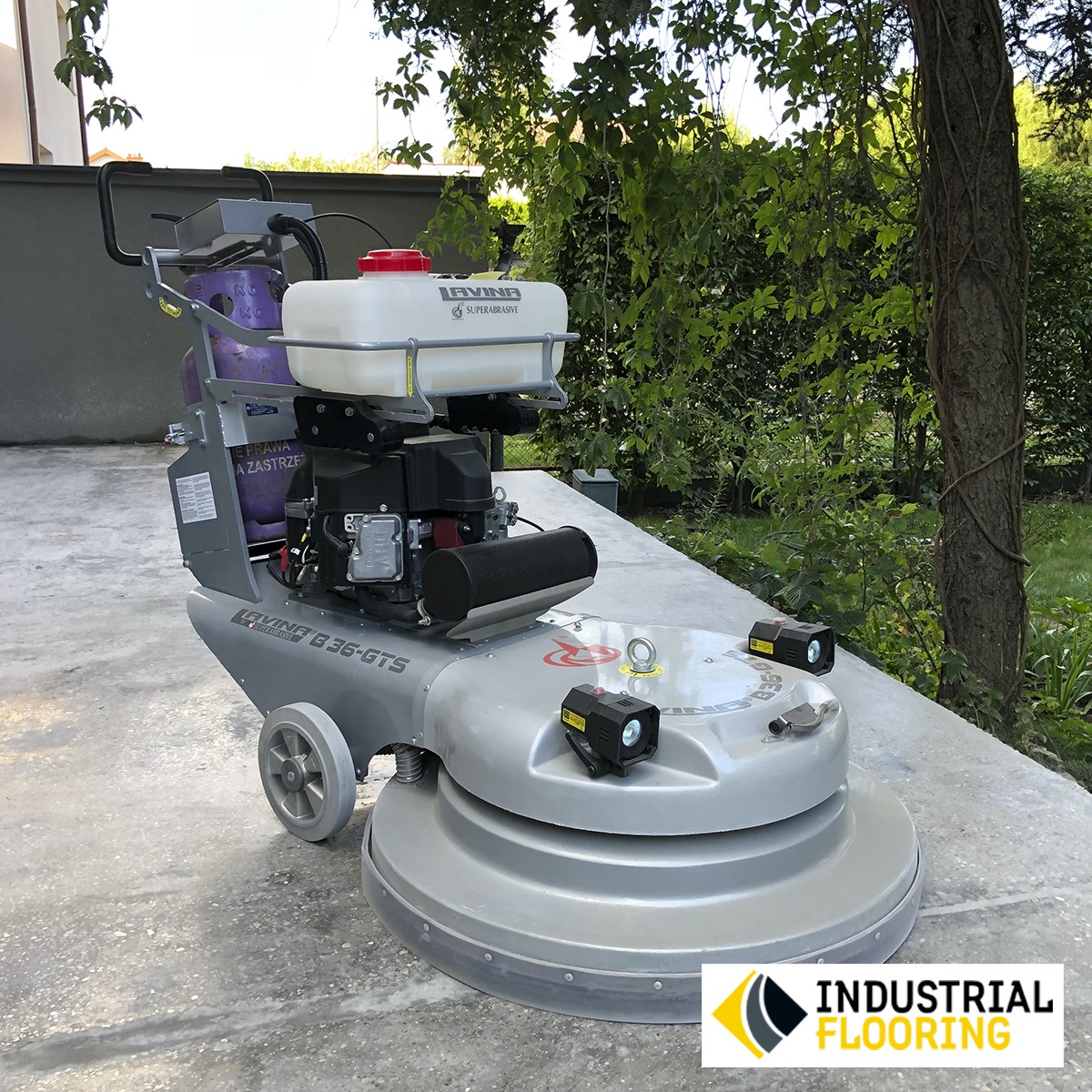 Szara szlifierka do posadzek marki Levig G 36-GTS z białym zbiornikiem na wodę, stojąca na szarej betonowej powierzchni na tle zieleni.