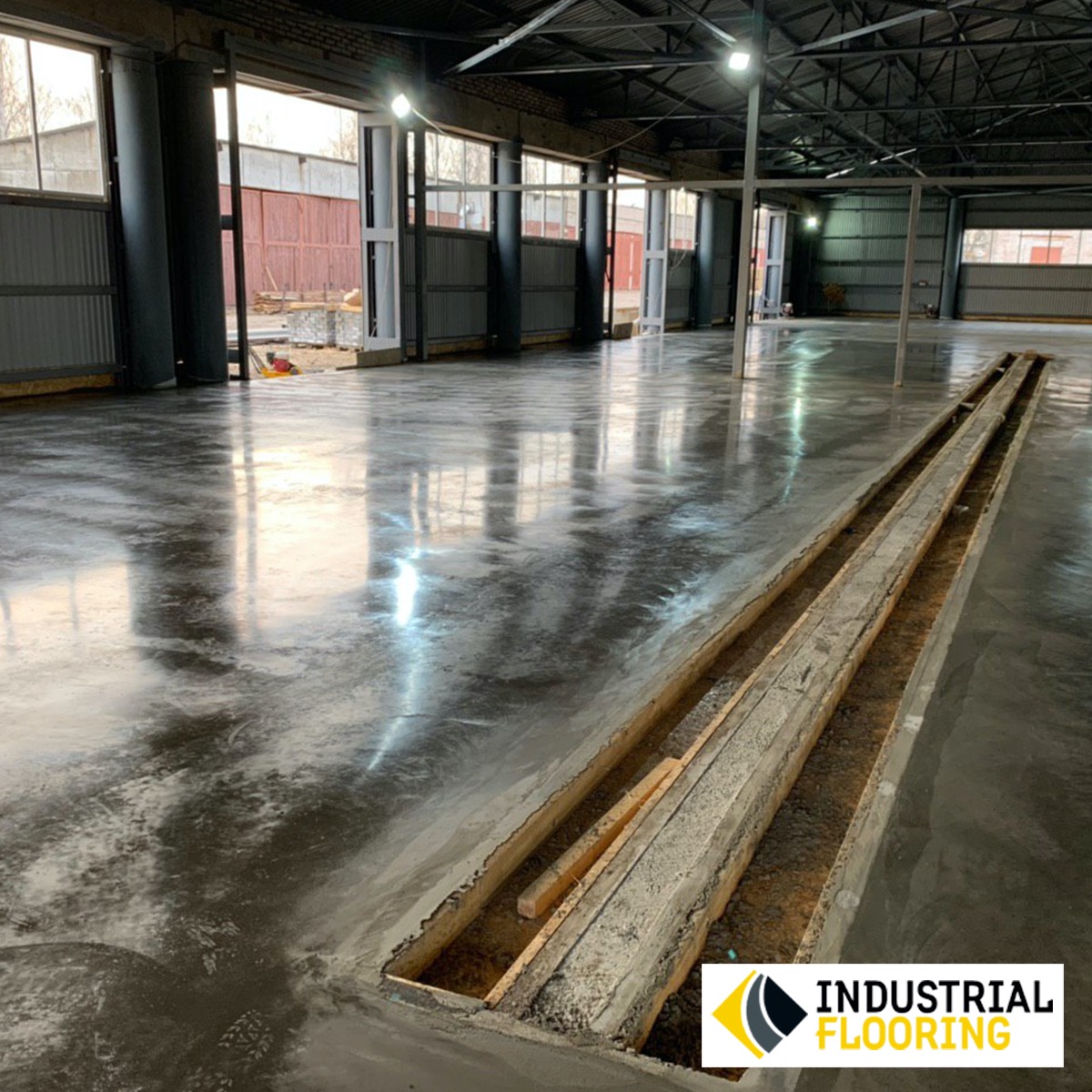 Błyszcząca, szara posadzka żywiczna w hali przemysłowej, z widocznym kanałem technicznym zabezpieczonym drewnianymi belkami, i logo w prawym dolnym rogu.