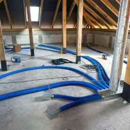 Niedokończona instalacja wentylacji na poddaszu: niebieskie, karbowane rury prowadzone po betonowej podłodze, narzędzia i skrzynka narzędziowa w tle, widoczne drewniane elementy konstrukcji dachu...