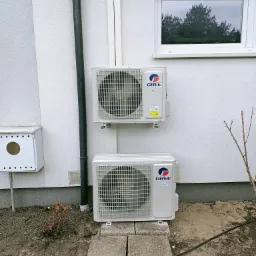 Dwa klimatyzatory marki Gree zamontowane na zewnętrznej ścianie budynku, połączone pionową rurą, widoczne betonowe płyty pod dolnym urządzeniem i okno z odbiciem drzew