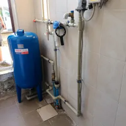 Instalacja hydroforowa w pomieszczeniu z beżowymi płytkami, widoczny niebieski zbiornik przeponowy Wimest ZPL/150/6, filtry wody z czarnym kluczem do odkręcania obudowy, orurowanie z tworzywa...