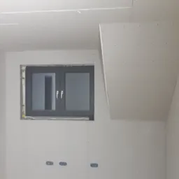 Poddasze z oknem w trakcie zabudowy płytami gipsowo-kartonowymi, widoczne kable elektryczne i otwory na gniazdka.