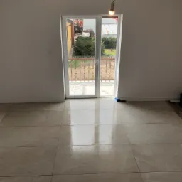 Beżowe, błyszczące płytki podłogowe w nowym pomieszczeniu z widokiem na ogród przez balkonowe drzwi, surowa żarówka zwisa z sufitu.