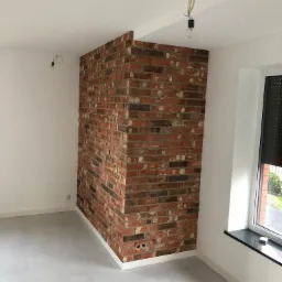 Wnętrze z fragmentem ściany wykończonym dekoracyjną cegłą klinkierową w odcieniach czerwieni i brązu, szara podłoga z płytek, białe ściany i sufit, okno z czarnym parapetem, wisząca żarówka bez...