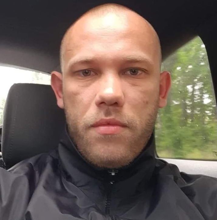 Portret mężczyzny z krótką brodą i ogoloną głową, ubranego w czarną kurtkę, siedzącego w samochodzie na tle rozmytej zieleni za oknem.