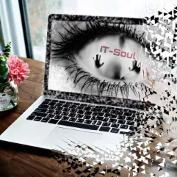 Laptop z wyświetlonym graficznym okiem i napisem IT-Soul, efekt rozpadu na trójkąty, obok filiżanka kawy i wazonik z kwiatami na drewnianym stole.