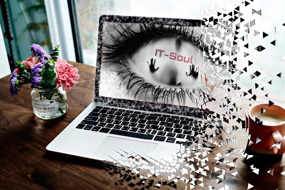 Laptop z wyświetlonym graficznym okiem i napisem IT-Soul, efekt rozpadu na trójkąty, obok filiżanka kawy i wazonik z kwiatami na drewnianym stole.