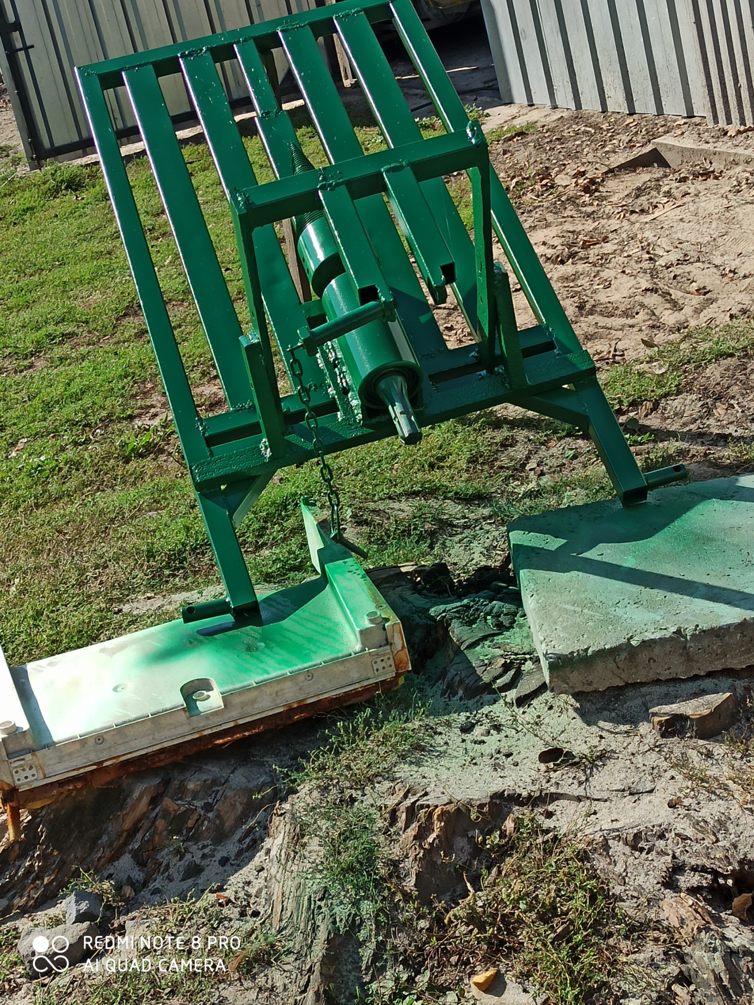 Świeżo pomalowany na zielono stojak z cylindrem hydraulicznym, obok leży element konstrukcyjny, również pomalowany na zielono, na zewnątrz, na trawie i piasku.