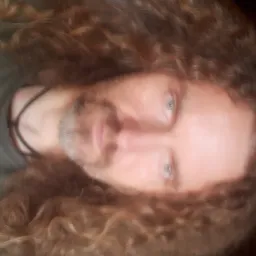 Portret mężczyzny z długimi kręconymi włosami i lekkim zarostem, skierowany w dół, z widocznymi niebieskimi oczami i ciemnym tłem.