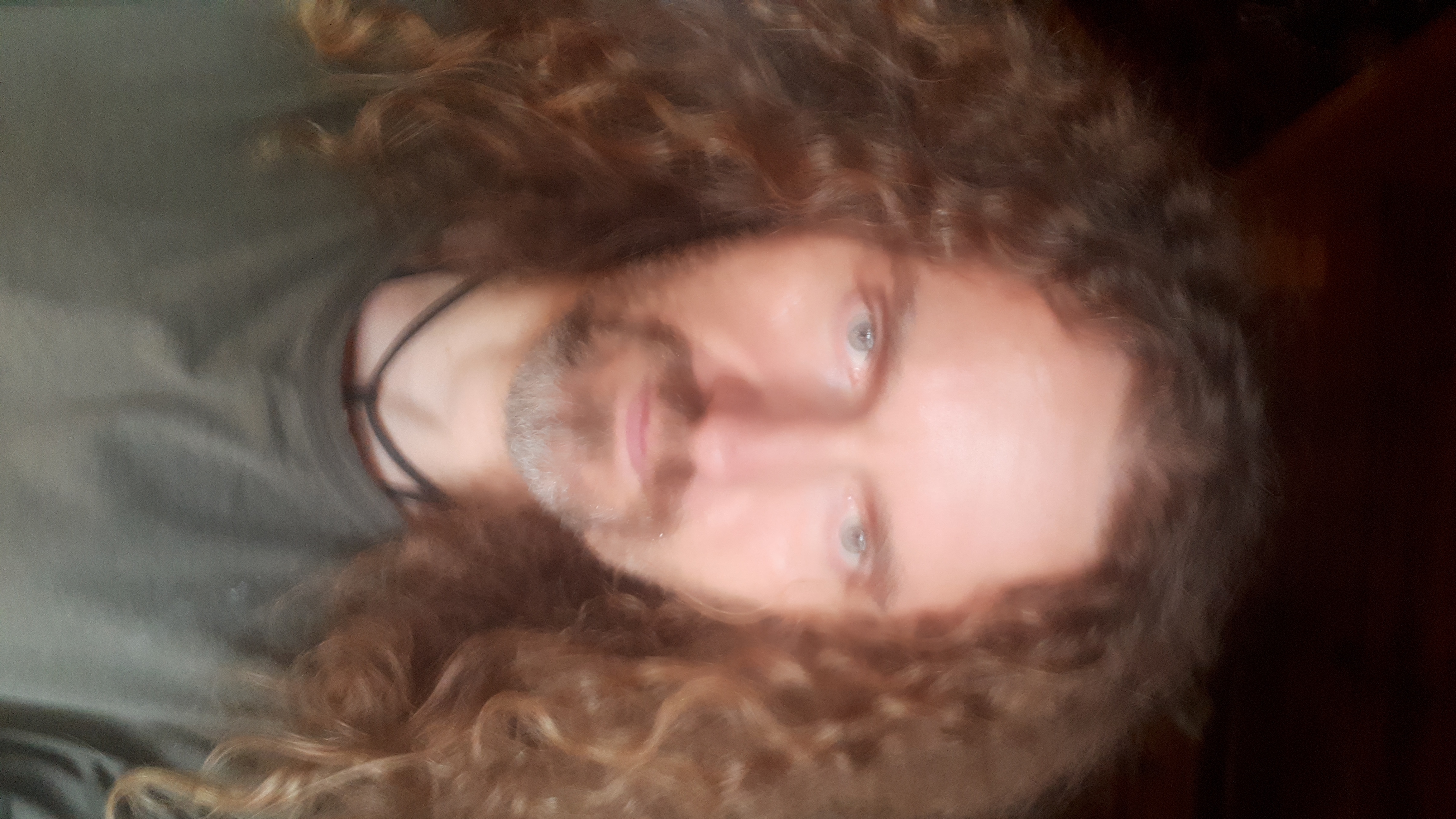 Portret mężczyzny z długimi kręconymi włosami i lekkim zarostem, skierowany w dół, z widocznymi niebieskimi oczami i ciemnym tłem.