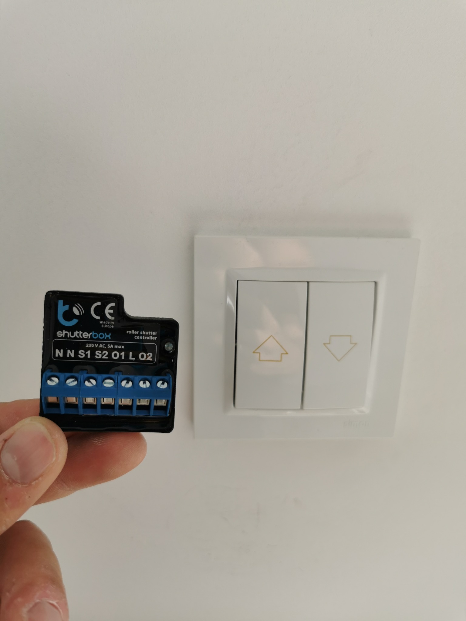 Czarny moduł sterujący roletami Shutterbox trzymany w dłoni, obok białego podwójnego włącznika ściennego z symbolami strzałek góra/dół, na tle białej ściany.
