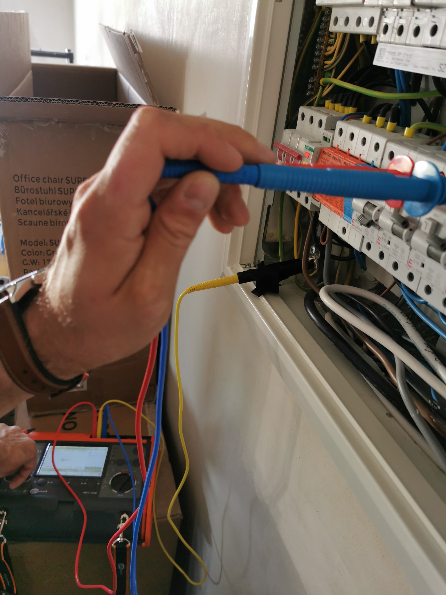 Elektryk mierzy parametry instalacji elektrycznej w rozdzielnicy za pomocą miernika wielofunkcyjnego, widoczne przewody pomiarowe i elementy zabezpieczeń