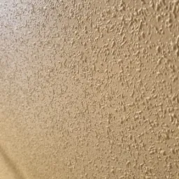 Beżowa ściana z wyraźną fakturą baranka, oświetlona z boku, ukazująca detale wykończenia. Widoczny fragment rury przy ścianie.