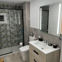 Krzysztof Piechota Usługi Malarskie dla Budownictwa - Nowoczesna łazienka po remoncie: szara glazura, kabina z geometrycznym wzorem, podświetlane lustro i drewnopodobna szafka pod umywalką. Minimalistyczny design.