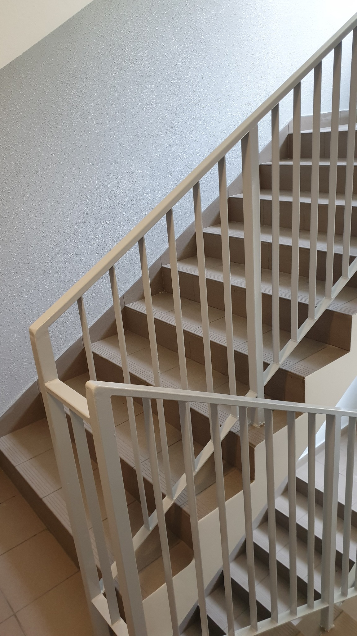 Klatka schodowa z jasnymi, metalowymi barierkami i schodami wyłożonymi płytkami. Ściana w tle ma delikatną, teksturowaną powierzchnię.