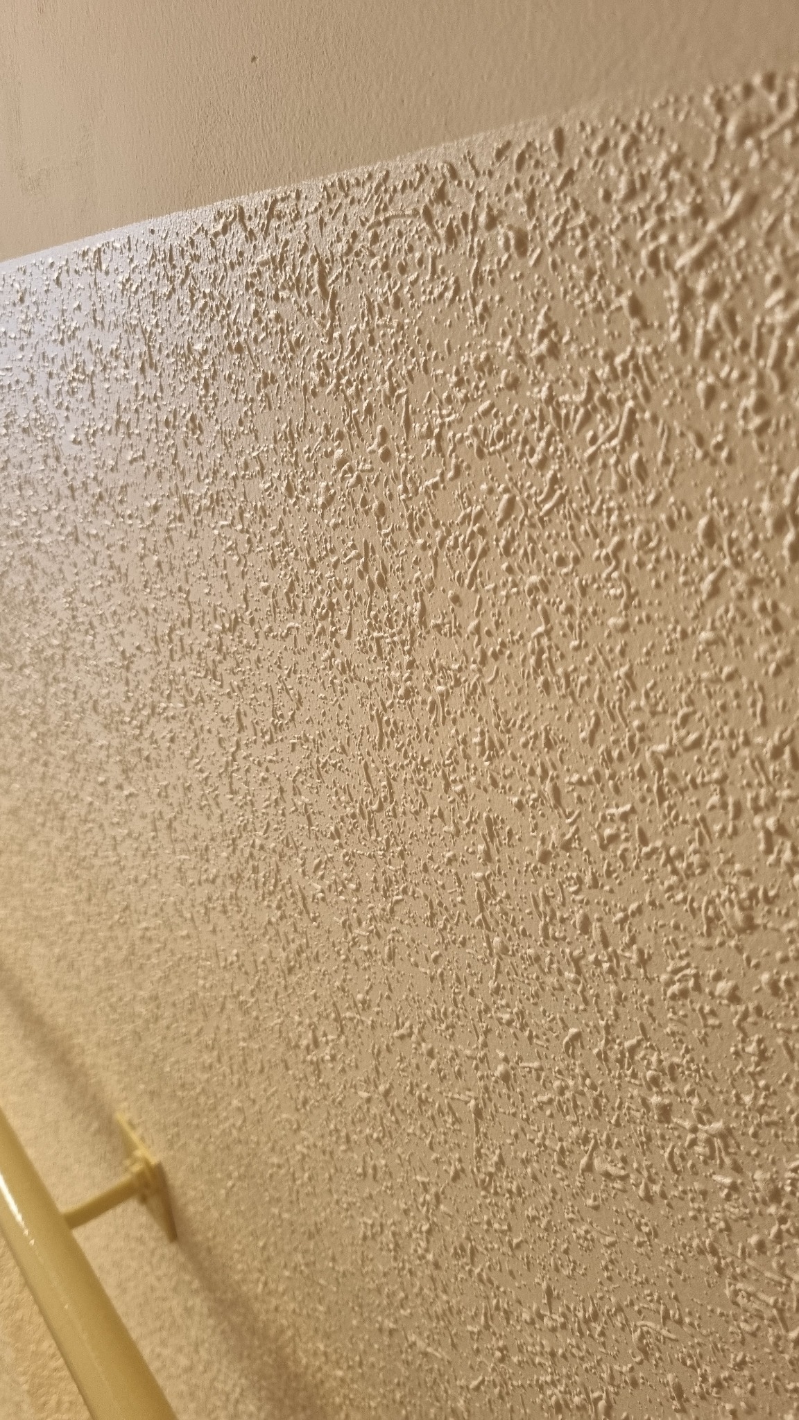Beżowa ściana z wyraźną fakturą baranka, oświetlona z boku, ukazująca detale wykończenia. Widoczny fragment rury przy ścianie.