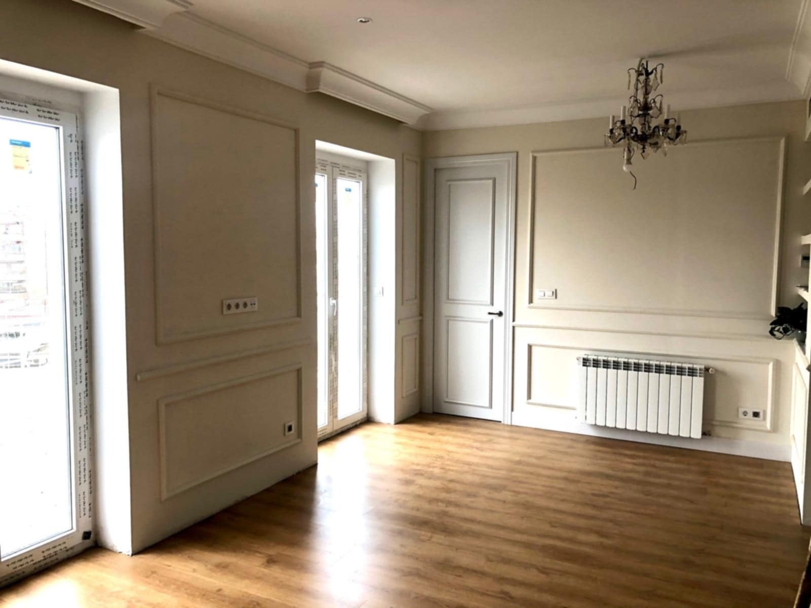 Jasne wnętrze po renowacji: parkiet, kremowe ściany z dekoracyjnymi panelami, białe drzwi i okna. Klasyczny żyrandol i grzejnik dodają elegancji.