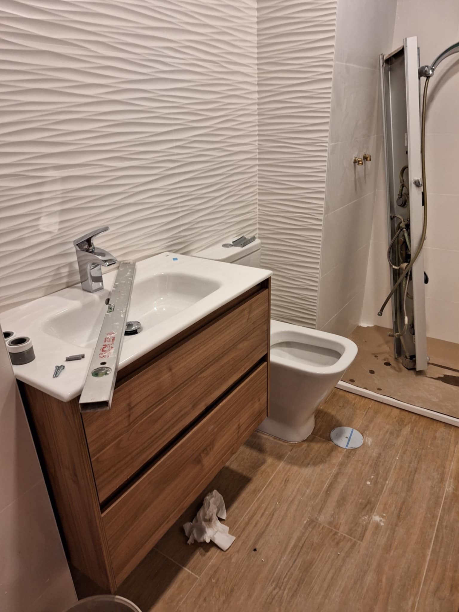 Montaż umywalki z drewnianą szafką w łazience z falistą płytką ścienną i częściowo zamontowaną kabiną prysznicową. Widoczna poziomica i elementy montażowe.