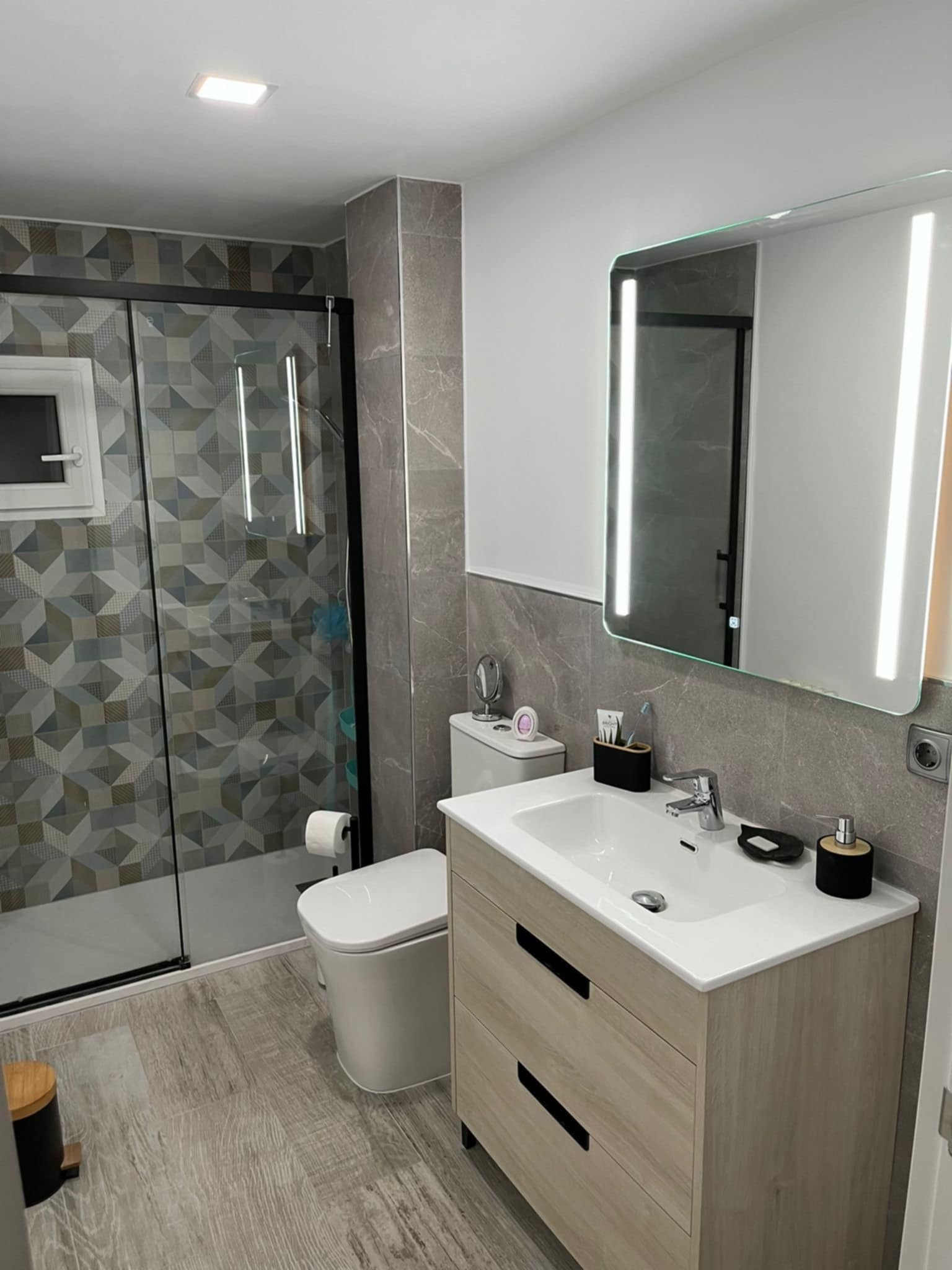 Nowoczesna łazienka po remoncie: szara glazura, kabina z geometrycznym wzorem, podświetlane lustro i drewnopodobna szafka pod umywalką. Minimalistyczny design.