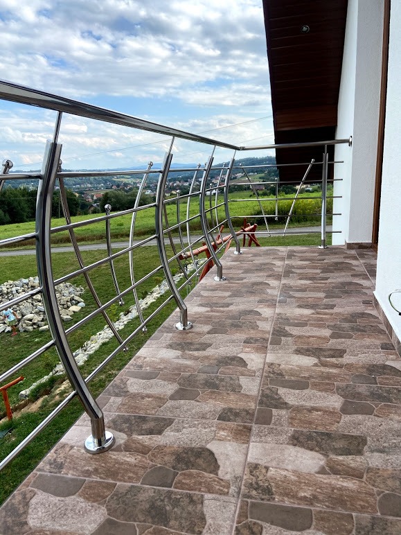 Balkon z nowoczesną, giętą balustradą ze stali nierdzewnej z widokiem na zielony krajobraz i zabudowania w oddali.