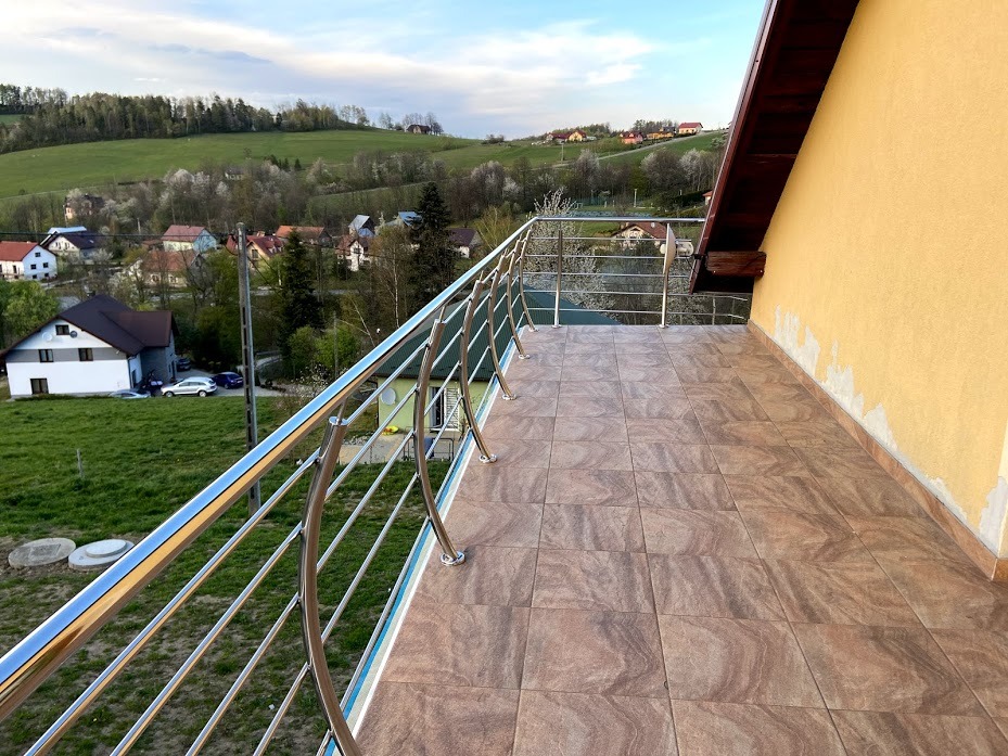 Balkon z nierdzewną balustradą z poziomymi prętami, widok na pagórkowaty krajobraz z zabudowaniami mieszkalnymi w oddali, płytki ceramiczne na podłodze balkonu imitujące drewno.