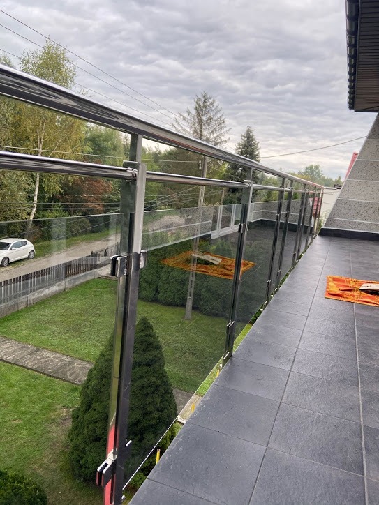 Szklana balustrada z nierdzewnymi elementami mocującymi na balkonie z szarą podłogą, widok na zielony ogród i ulicę w tle, pochmurne niebo.