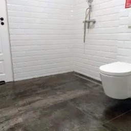 Nowoczesna łazienka z wiszącą toaletą, białą armaturą i płytkami imitującymi cegłę oraz ciemną podłogą z płytek w odcieniach szarości i brązu.