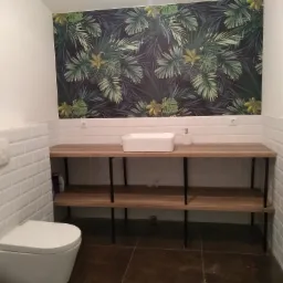 Biała toaleta z deską sedesową, umywalka nablatowa na drewnianym blacie z czarnymi nogami, białe kafelki typu metro i tapeta w tropikalne liście w łazience.