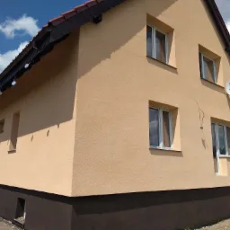 Dom jednorodzinny z beżową elewacją i ciemnobrązową podmurówką, widoczne okna z białymi ramami, antena satelitarna i ozdoby z płyt CD pod dachem.