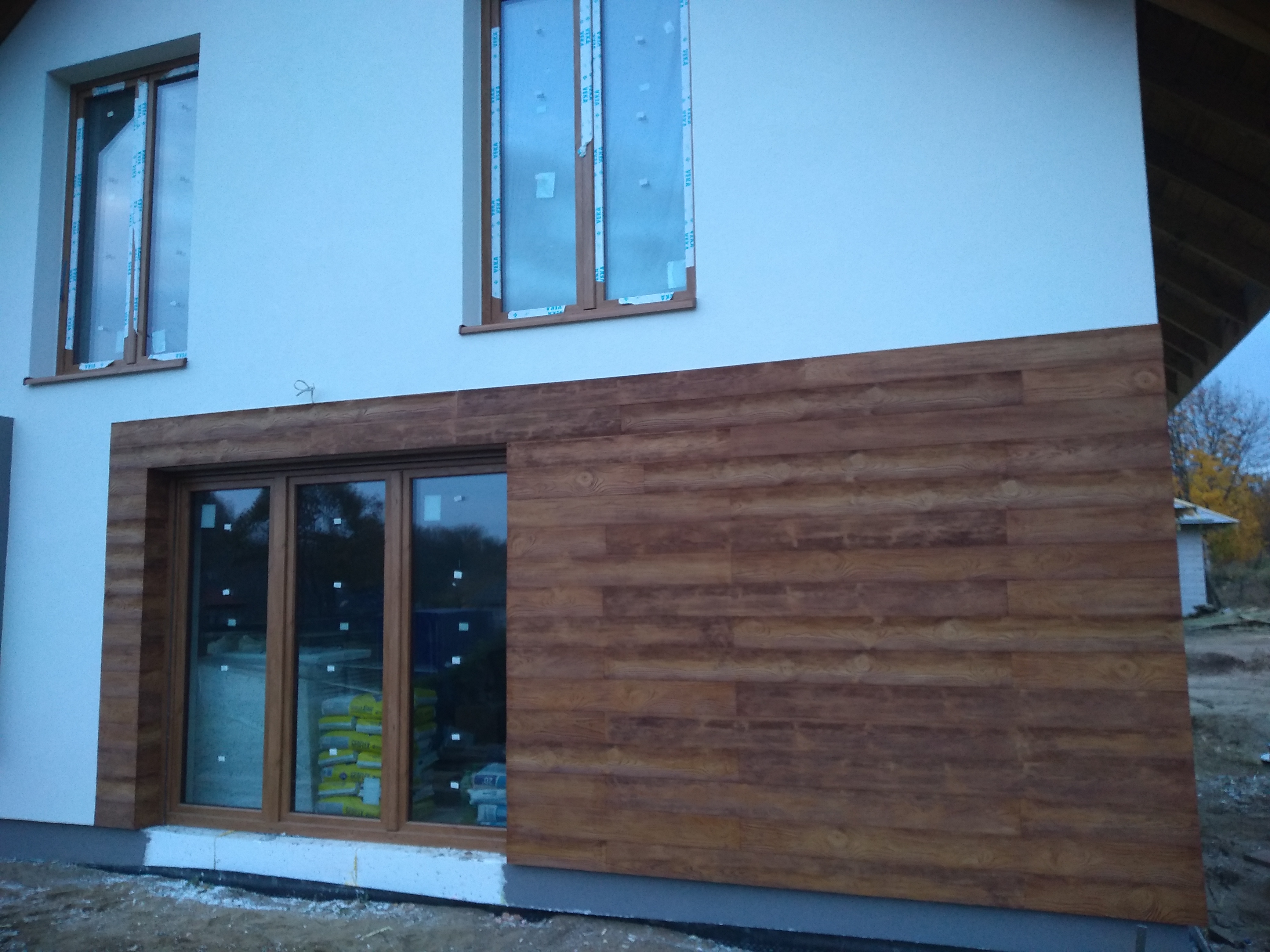 Fragment domu z drewnopodobną elewacją, widoczne okna z folią ochronną i worki z materiałami budowlanymi wewnątrz.