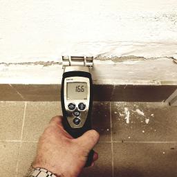 Drymaster - Pomiar wilgotności ściany przy pomocy wilgotnościomierza Testo 616 wskazującego 16.6%, widoczne ślady zawilgocenia na ścianie i odpadający tynk, fragment ręki z metalowym zegarkiem.