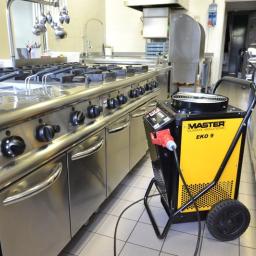 Drymaster - Przemysłowy osuszacz powietrza Master EKO 9 na kółkach, ustawiony w kuchni z nierdzewnymi urządzeniami, podłączony do gniazdka z czerwoną wtyczką.