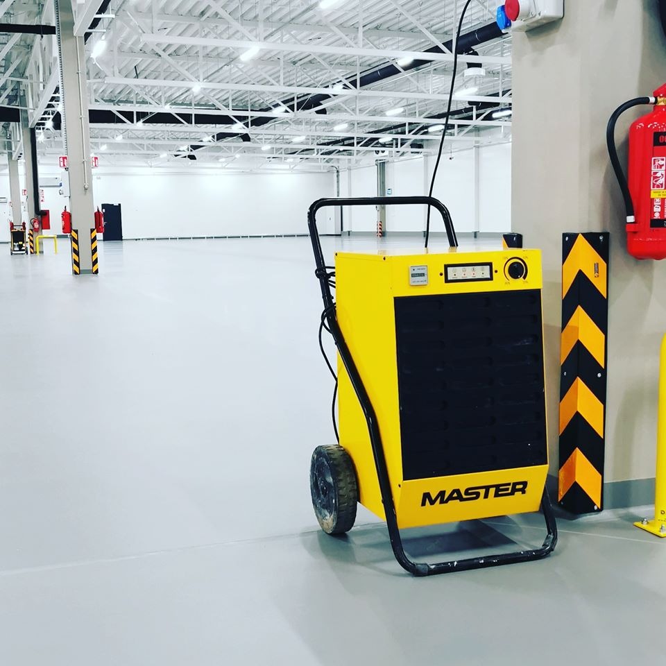 Żółty przemysłowy osuszacz powietrza marki Master z czarnym panelem na kółkach, stojący na szarej posadzce w dużym, jasnym, pustym magazynie z białą konstrukcją dachu