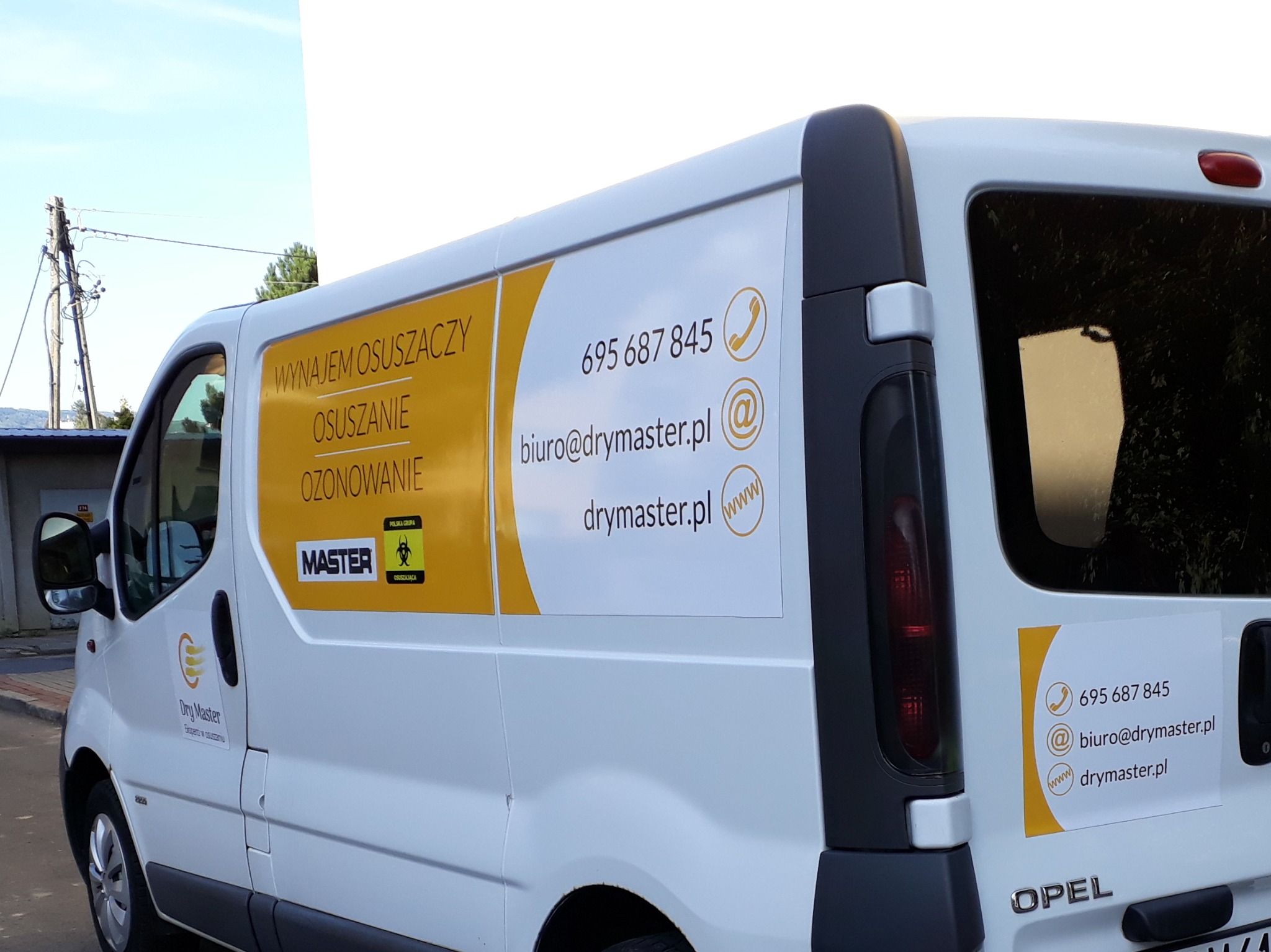 Biały samochód dostawczy firmy Dry Master z reklamą usług wynajmu osuszaczy, osuszania i ozonowania oraz danymi kontaktowymi: numer telefonu, adres e-mail i strona internetowa.