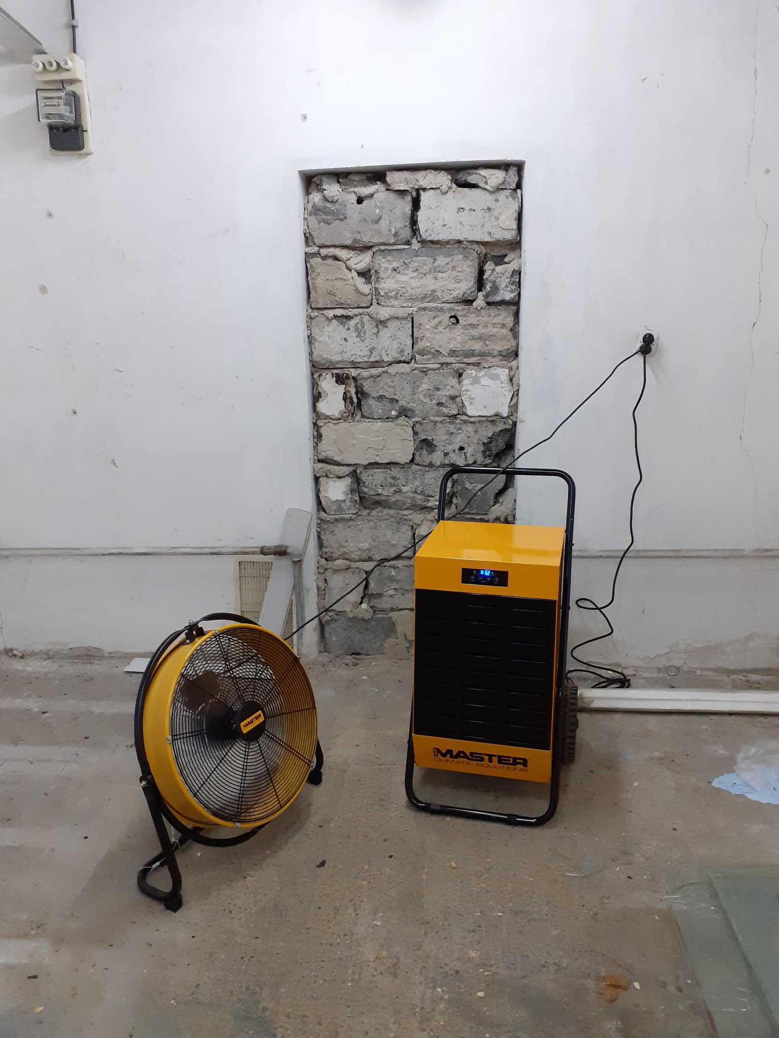 Dwa żółte urządzenia Master: wentylator osuszający i nagrzewnica stojące w surowym pomieszczeniu z odsłoniętą cegłą w otworze ściennym, widoczne ślady wilgoci na ścianach.