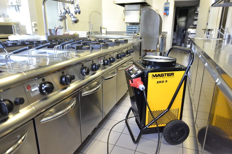 Przemysłowy osuszacz powietrza Master EKO 9 na kółkach, ustawiony w kuchni z nierdzewnymi urządzeniami, podłączony do gniazdka z czerwoną wtyczką.