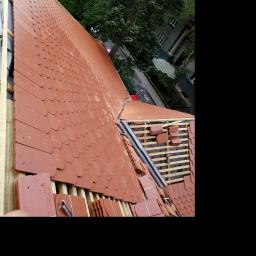 Usługi Dekarskie - Montaż czerwonej dachówki karpiówki na drewnianej konstrukcji dachu, widoczne elementy mocujące i częściowo ukończona połać dachu, w tle zieleń drzew i budynek.