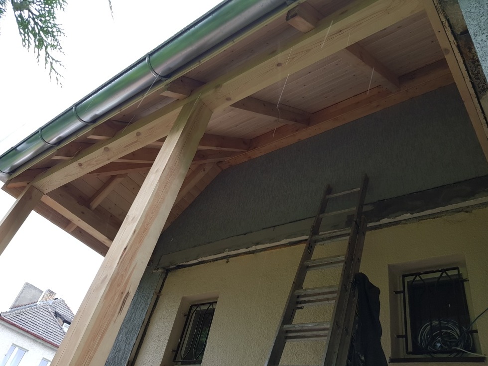 Konstrukcja drewniana z zamontowaną rynną, drabina oparta o ścianę budynku z widocznymi oknami w trakcie prac wykończeniowych.