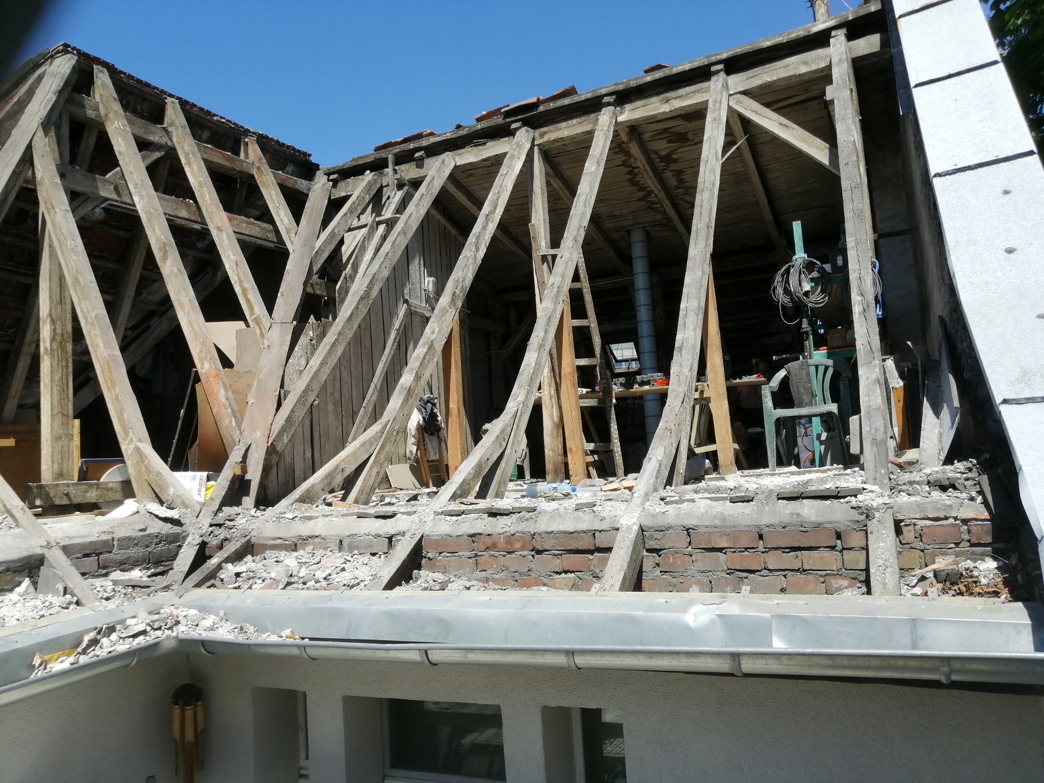 Demontaż starego dachu z widoczną konstrukcją drewnianą, ceglanym kominem i fragmentem zamontowanej rynny.