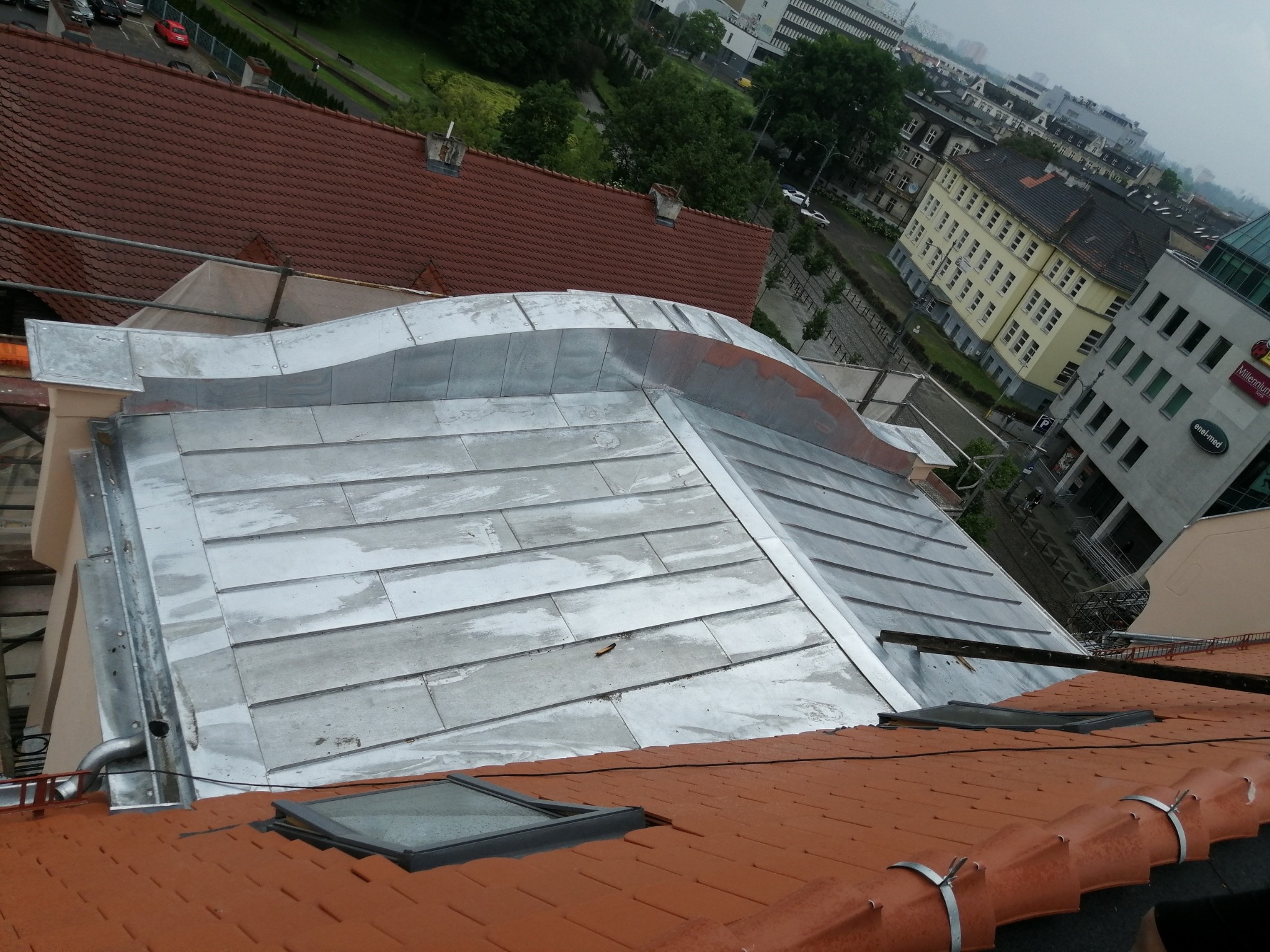 Widok z góry na fragment dachu z blachy trapezowej, obróbki blacharskie przy krawędzi, okno dachowe i fragment dachu z dachówki ceramicznej w kolorze ceglastym, na tle zabudowy miejskiej.